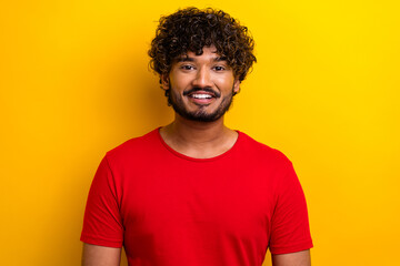 Smiling young man in a bright red t-shirt standing against a cheerful yellow background, radiating positivity with curly black hair.