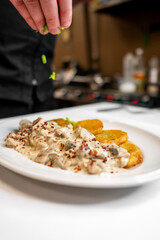 A chef sprinkles fresh herbs over a creamy mushroom dish served with golden fried plantains on a white plate, showcasing culinary skills in a vibrant kitchen setting.