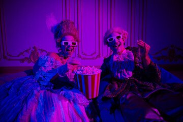 A joyful couple dressed in elegant baroque costumes enjoys popcorn indoors, illuminated by neon purple light