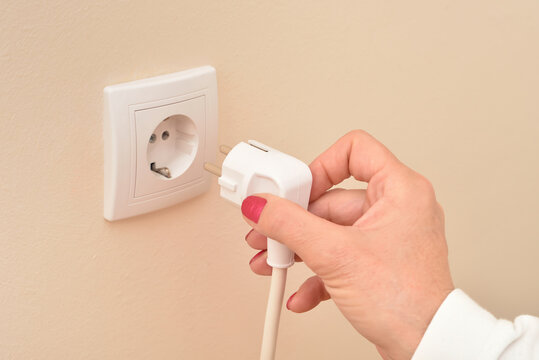 Hand of a woman holding white electric cable next to the white power socket on the pale orange wall. Copy space. Close-up. Concept of electricity consumption.   