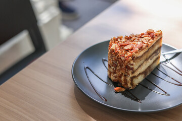 A slice of carrot cake with walnut toppings on the black plate and chocolate strips as decoration. Placed on the cafe wooden table. Empty space for text and messages