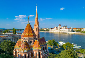 Fototapeta premium View of Budapest along the Danube River, in the foreground the Reformed Church with the patterned roof tiles and tall spire, in the background the Hungarian Parliament Building