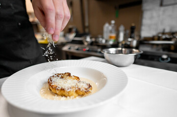 A chef's hand sprinkles grated cheese over a beautifully plated dish in a modern kitchen. The circular plate features a gourmet preparation, showcasing attention to detail and culinary artistry.