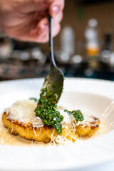 A close-up shot of a hand using a spoon to spread green sauce over a plate of freshly cooked food topped with grated cheese. The setting showcases a busy kitchen in the background.