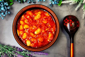 Bean soup, beans in tomato in a clay plate, wooden spoon, berries and herbs on a gray background