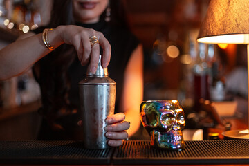Girl bartender prepares delicious alcoholic cocktail in a glass skull on the bar counter. closes the shaker