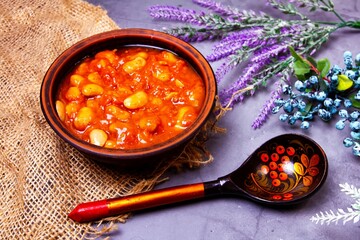 Bean soup, beans in tomato in a clay plate, wooden spoon, burlap, berries and herbs on a purple background