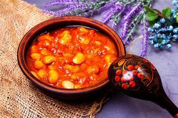 Bean soup, beans in tomato in a clay plate, wooden spoon, burlap, berries and herbs on a purple background