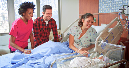 Female Nurse Wearing Scrubs Meeting Parents With Newborn Baby Boy Lying In Hospital Bed And Cot