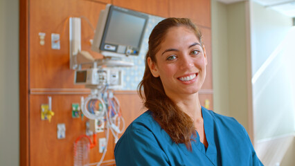 Portrait Of Female Doctor Or Nurse Wearing Scrubs At Work Standing Beside Hospital Equipment