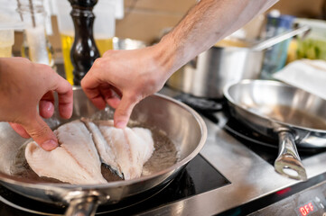 Two hands carefully place fish fillets in a hot frying pan, cooking in a bustling kitchen. The scene captures the culinary process, showcasing fresh ingredients and a vibrant kitchen atmosphere.