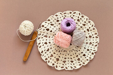 Top view of skeins of light natural cotton yarn for crocheting and crochet hook on handmade lace round doily on mocha color background. Crochet as a hobby. DIY concept. Beautiful still life. Close-up