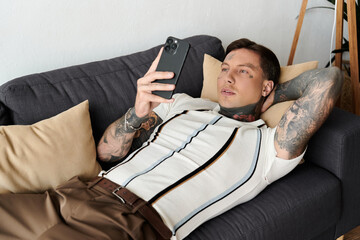 Man with tattoos lounges casually on a couch while checking his smartphone in a cozy room.