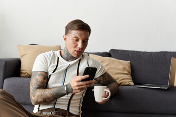 Tattooed young man sits on couch, focused on smartphone and sipping from a cup in casual setting.