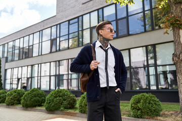A young handsome man with tattoos poses calmly outside a contemporary building during the day.