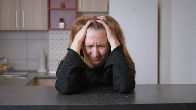 Young woman sits alone at kitchen counter, resting her head in her hands. Sadness and despair fill her expression, reflecting deep emotional struggle and loneliness