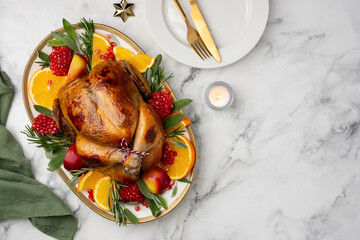 Christmas roasted chicken with apples, oranges, pomegranates and herbs on a large plate on a marble background. Festive poultry dish. Top view. Copy space