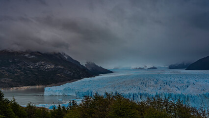 The endless blue glacier stretches between the mountains to the horizon. A mass of ice with cracks, sharp peaks, and crevices. Thawed ice floes float in the lake. The clouds. Fog. Green vegetation