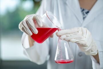 A scientist wearing gloves is pouring a red liquid from a beaker into an Erlenmeyer flask.