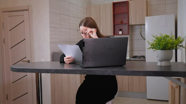 Female accountant with glasses reading documents and wondering with glasses on her forehead while working on laptop at her desk, showing difficulties in managing documents