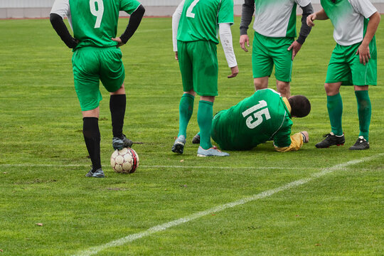A foul in European football, a player injury during a match, a free kick awarded after the foul. A footballer lies on the green grass field surrounded by teammates.