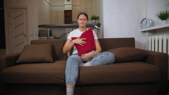 Woman suffering from strong abdominal pain while sitting on brown couch in her living room. She is clutching red pillow and wincing in discomfort