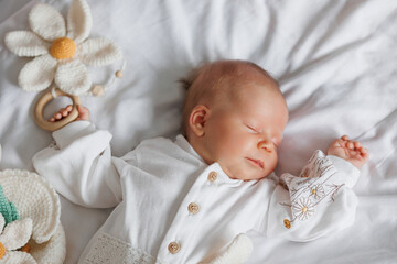 A baby is sleeping on a bed with a white blanket and a flower toy. The baby is wearing a white...