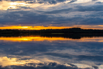 A serene sunset over a calm lake with dramatic clouds, golden and orange reflections on the water, and a silhouette of distant trees.