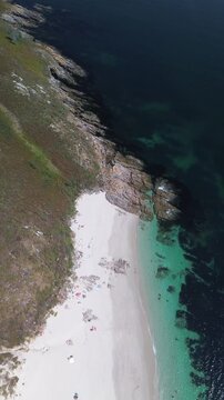 Aerial view of Melide Beach on Ons Island on sunny summer morning. Camera moves backward, revealing lush dunes, white sand, turquoise waters, and scattered rocks, with tourists enjoying the beach.