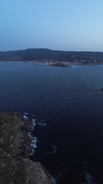 Blue hour aerial descent over the rocky, shrub-covered cliffs of Monteferro. The waves of the calm Atlantic Ocean gently crash aganist the cliffs, with Baiona's parador and harbor in the distance.