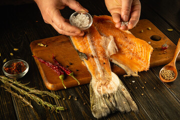 An artisan chef skillfully seasons a fresh salmon fillet on a wooden cutting board. Surrounding the...