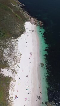 Aerial view of Melide Beach on Ons Island during a sunny summer morning. Camera moves forward, capturing the white sand, turquoise waters, rugged rocks, and a scenic dirt trail next to the dunes.