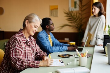 Side view of mature female employee writing in calendar table planning monthly tasks while sitting at working desk in office, copy space