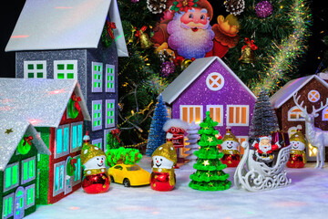Among five decorated houses with bright lights in the windows, a small green Christmas tree is surrounded by Christmas decorations in the shape of a snowman and Santa Claus on a sleigh