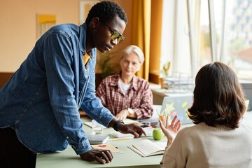 Side view of fashionable Black man in denim shirt outfit for office styled with colorful yellow glasses chatting with female colleague, while standing next to desk at workplace