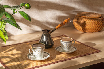 Two cups of coffee and cezve on the table in the patio lit by morning sun.	