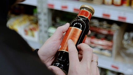 A customer in black studies the label on a bottle of sauce. Man carefully reads a product label in a busy supermarket aisle, making a healthy choice based on nutrition and ingredients