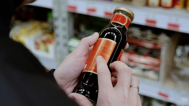 A customer in black studies the label on a bottle of sauce. Man carefully reads a product label in a busy supermarket aisle, making a healthy choice based on nutrition and ingredients