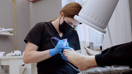Podiatrist wearing black mask and blue gloves using electric nail drill for pedicure treatment on...