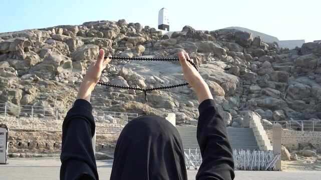 A girl in a black hijab holds a rosary with a crescent moon in her hands against the backdrop of the sacred Mount Arafat. Space for text.