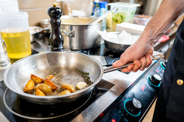 A chef's hand holds a frying pan on a stove, cooking golden-brown potatoes with fresh herbs. The kitchen is bustling with various ingredients and utensils, reflecting a lively cooking environment.