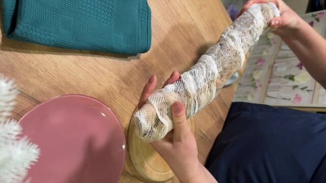 A woman is removing the cheesecloth that the cured pork is wrapped in. She makes a loin, dried pork tenderloin, processing pork at home. Next to it is a branch of artificial pine. Vertical video.