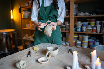 Obraz premium Hands of unrecognizable woman artisan molding clay mug in pottery workshop. Manual labor unique ceramic pieces, shaping wet clay, preparing for drying and firing process in work studio class.