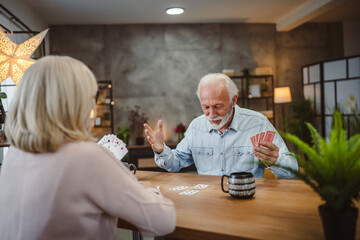 senior husband play cards with wife on wooden table at home