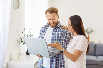 Young happy couple standing in cozy living room looking at laptop screen, watching video, buying goods, tickets online, using e-commerce app, feel satisfied, enjoy browsing web, planning leisure time