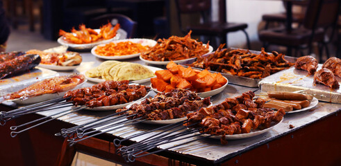 A vibrant display of grilled and roasted street food, featuring skewers of meat, roasted sweet potatoes, sausages, fish, shrimp, and flatbreads. Perfect for food photography, culinary promotions.