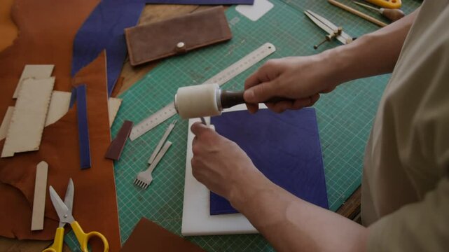 High angle shot of unrecognizable craftsman using mallet and chisel to work on dark blue leather surrounded by leather working tools, materials, and finished leather wallet in workshop