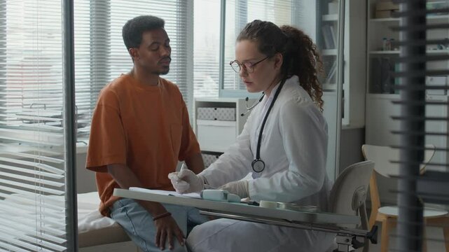 Medium wide shot of young Caucasian female doctor in white coat and sterile gloves examining young Black male patients eyes using diagnostic penlight then taking notes in outpatient card