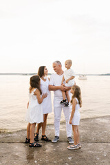 Happy family is standing on beach with boat in the background. Father is holding baby and other children are standing around him and mother