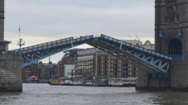 Tower Bridge in London with bascules raised for a passing boat, set against an overcast sky. Historic buildings, including Butler's Wharf, line the Thames.